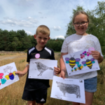 Photo of two young artists holding up their finished colouring sheets, with bees, birds and a Belted Galloway cow.