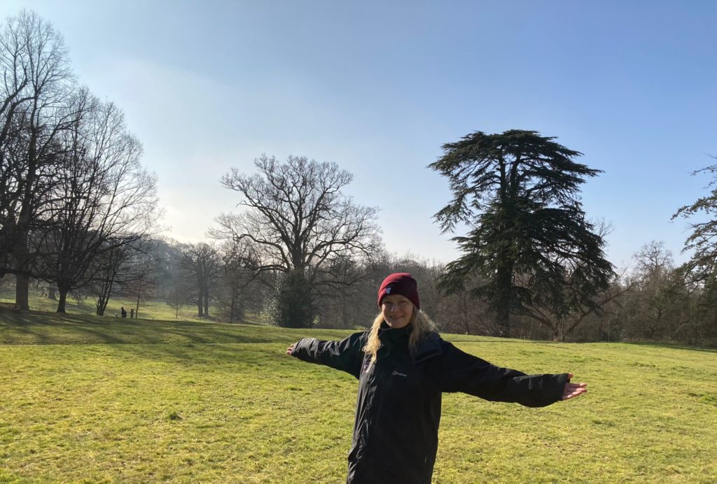 Photo of Warden Ruth with her arms outstretched, looking happy at Lily Hill park, amongst the trees!