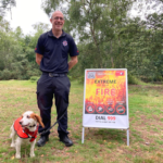 Photo of a man in a Fire Service uniform with a spaniel by his side. A sign says "Extreme risk of Fire".