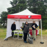 Photo of our team sheltering from the weather under a gazebo. The tables are all setup for colouring. Victoria stands in front with a litter picker and a bag of rubbish. Mike displays a leaflet about butterflies!