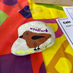 Colourful photo of a painted stone on a brightly coloured tablecloth. The stone has a brown bird painted on it and the word "Woodlark".