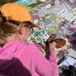 Photo of a young girl in an orange hat colouring in the outline of a snake. It's part of big, colourful artwork.
