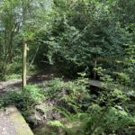 Photo shows a finger post pointing the way at a junction of paths beside a stream. A small footbridge spans the stream.