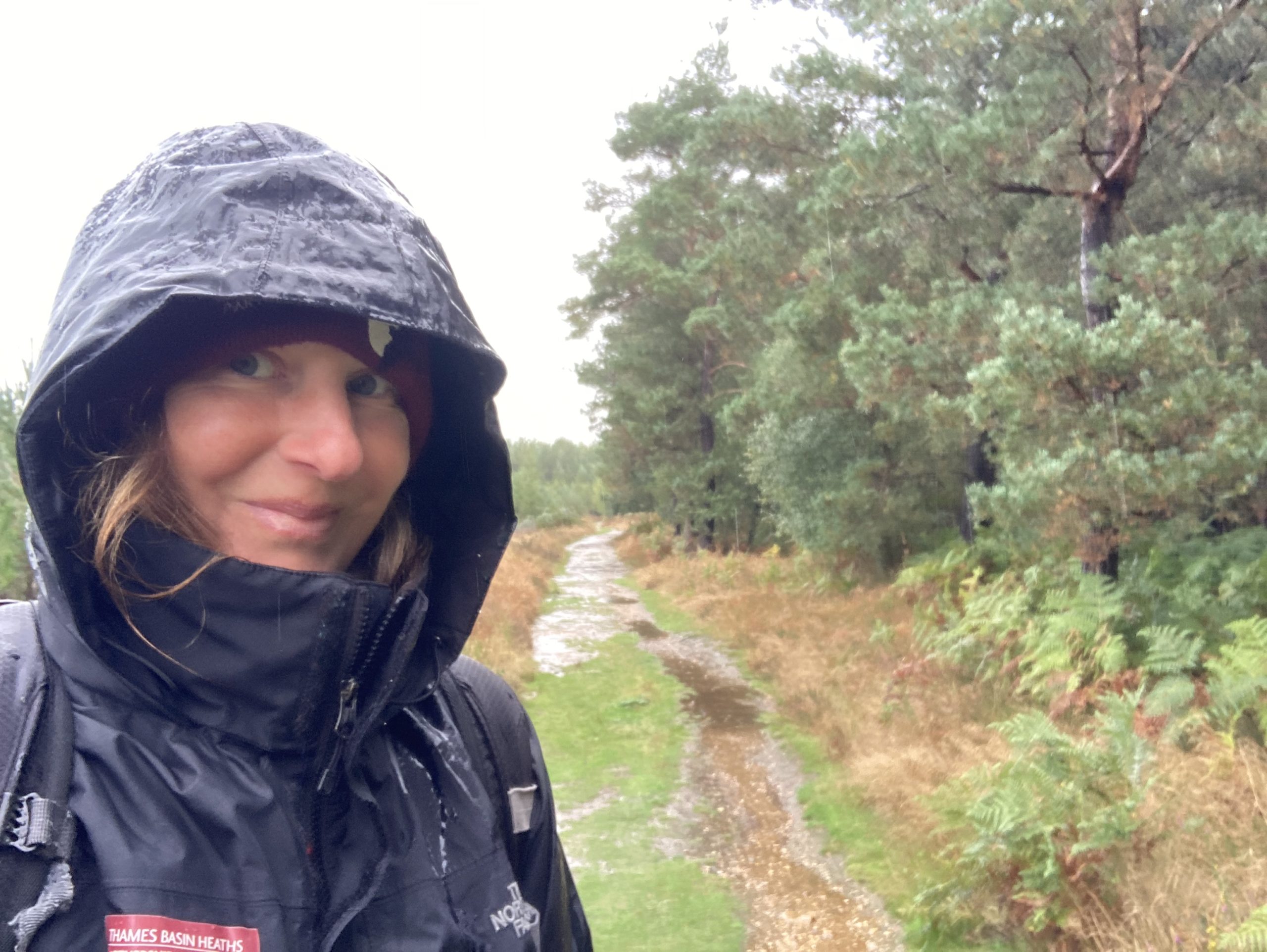 Photo of a Communications Officer Nicky in a forest, wearing a black waterproof coat with hood up, in the rain