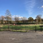 Photo of surfaced car park area boundaried by black metal fence, with grassy area and trees behind