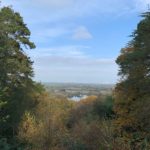 Photo shows a view out across Surrey. Tall trees frame the view and the colours of the leaves shows it's early autumn.