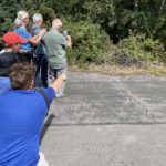 Photo of an old tarmac airfield with a group of adults and children looking at the scrubby edge and taking photos (there is an adder out of sight, amongst the brambles)