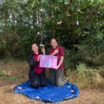 Two Thames Basin Heaths staff members kneel amongst the greenery holding a sign and pointing at decorative dragonflies which hang from the trees above them.