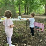 Two young children stand at a craft table amongst a woodland scene.